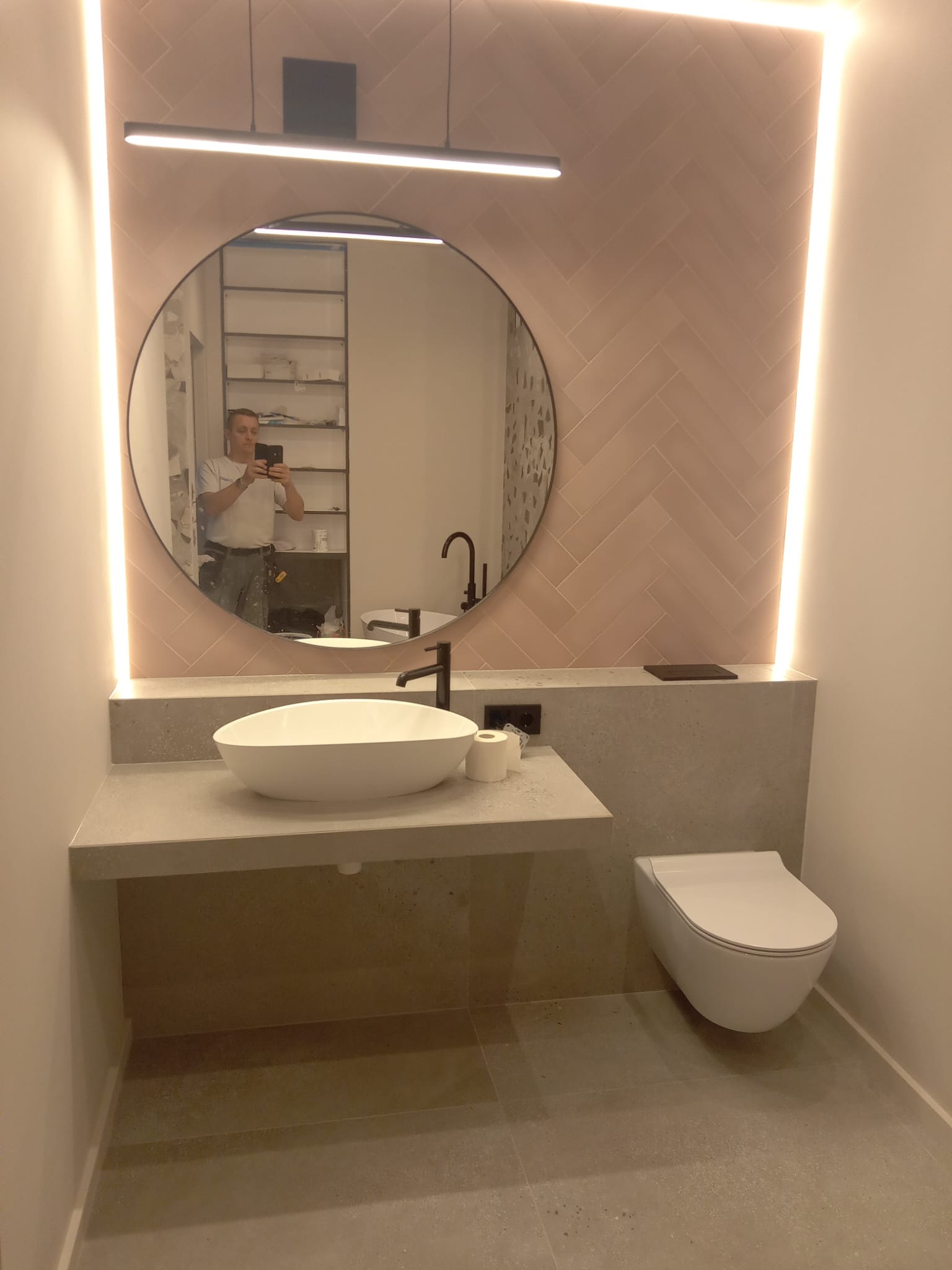 Nowoczesna łazienka z owalną umywalką nablatową, podwieszaną toaletą i różową ścianą w jodełkę, oświetlona ledowymi paskami. W lustrze odbija się osoba robiąca zdjęcie, widoczny również fragment...