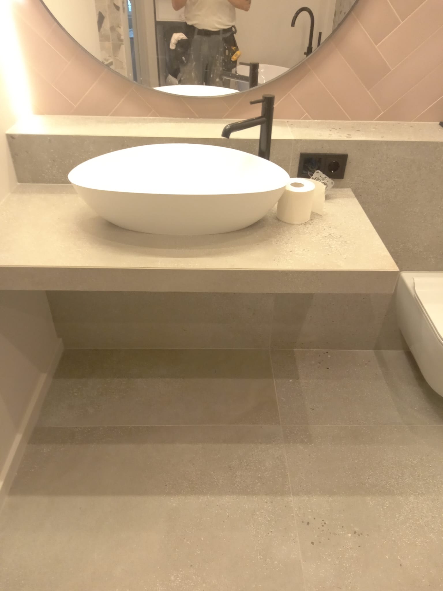 Nowoczesna łazienka z okrągłą umywalką nablatową, czarną armaturą i jasnoróżowymi płytkami ułożonymi w jodełkę nad blatem, widoczne odbicie osoby w lustrze.