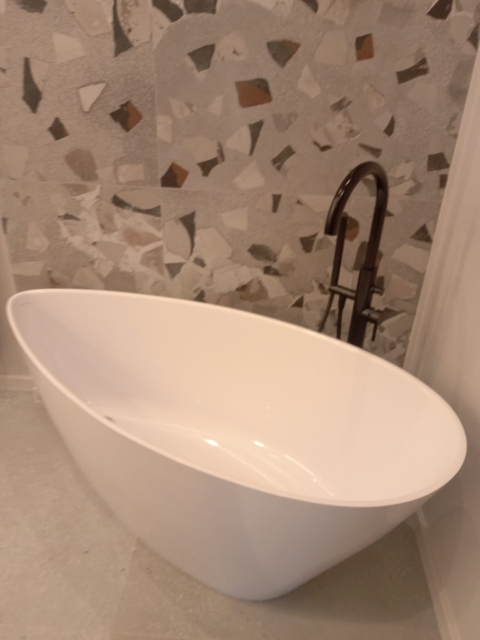 Wolnostojąca, owalna wanna z czarną armaturą na tle ściany wykończonej mozaiką z nieregularnych kamieni w odcieniach beżu, brązu i szarości.