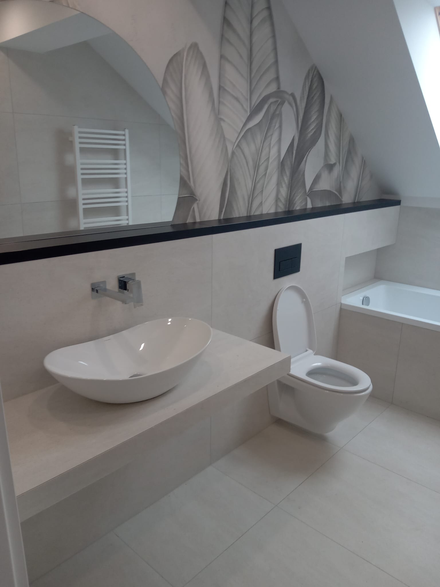 Nowoczesna łazienka na poddaszu z białą, owalną umywalką nablatową, wiszącą toaletą z czarnym przyciskiem, wanną w zabudowie i tapetą z motywem liści w odcieniach szarości.