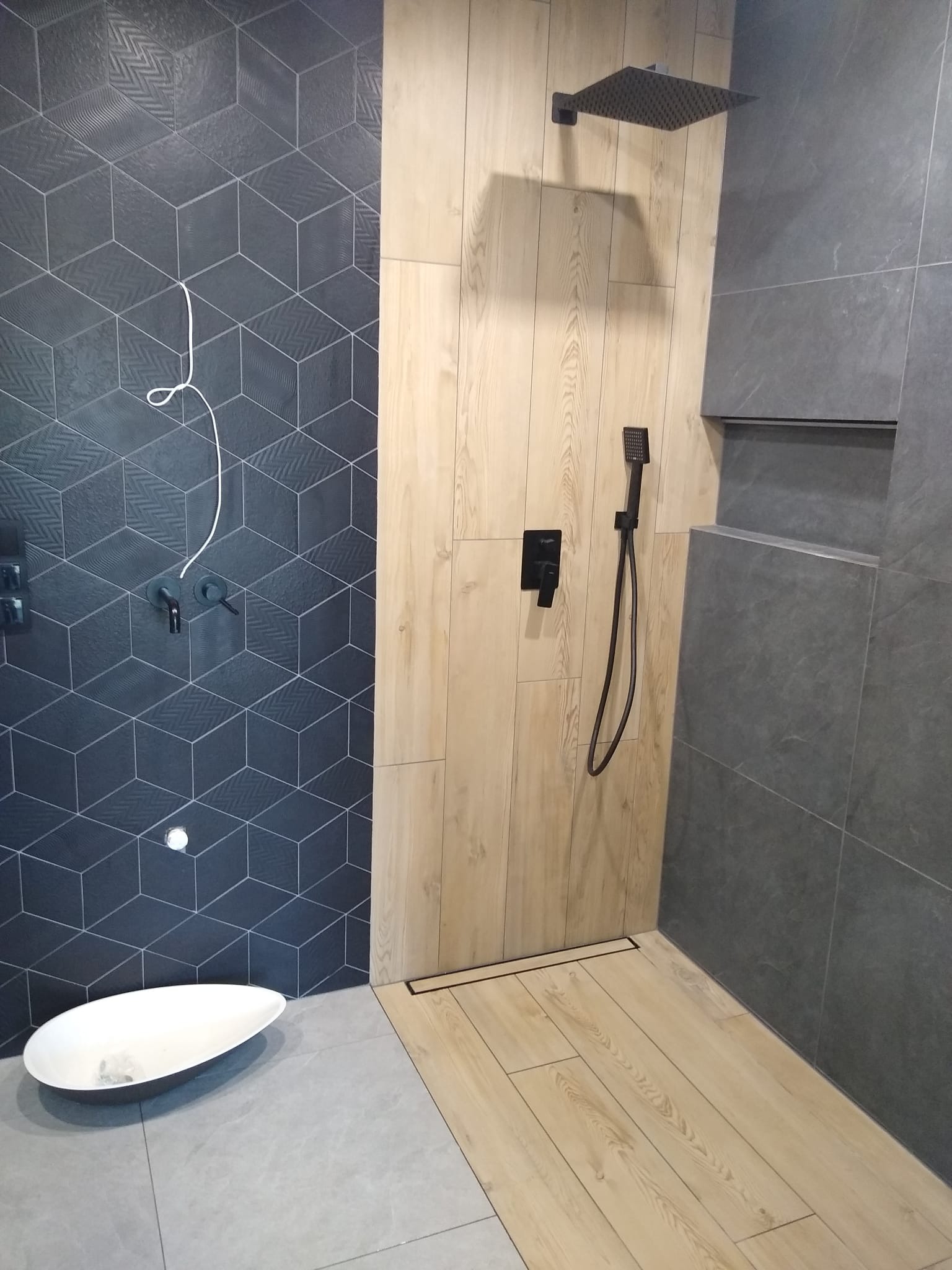 Nowoczesny prysznic z czarną armaturą, geometrycznymi płytkami w odcieniach szarości i drewnopodobną okładziną, widoczne niedokończone podłączenie instalacji.