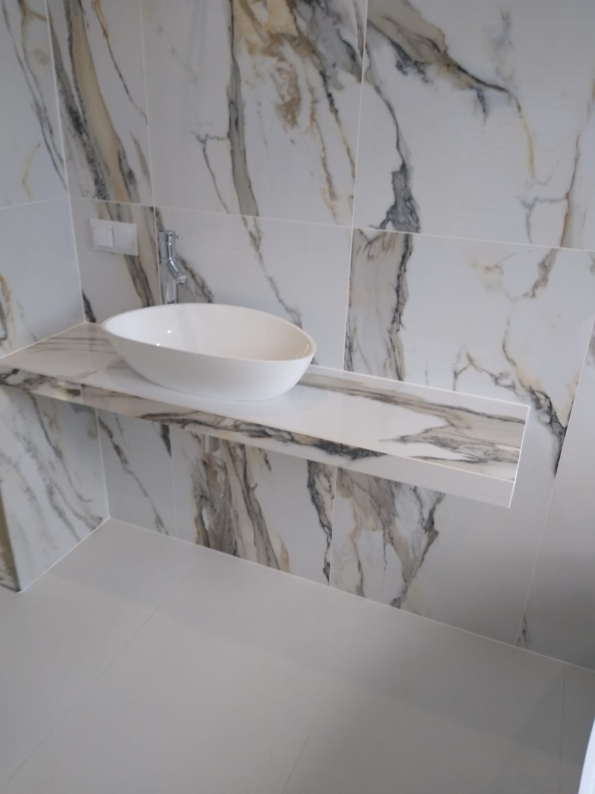 Biała, owalna umywalka nablatowa zamontowana na marmurowym blacie w łazience wykończonej płytkami imitującymi marmur z szarymi i złotymi żyłami.