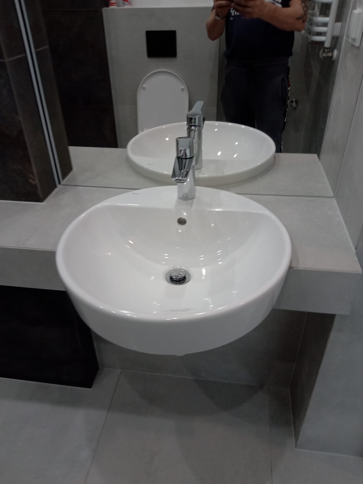 Nowoczesna, biała umywalka nablatowa z chromowaną baterią w szarej łazience z widocznym odbiciem osoby robiącej zdjęcie.