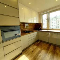 Jasna kuchnia na wymiar z drewnianym blatem i panelami, widok na budynek mieszkalny przez okno.