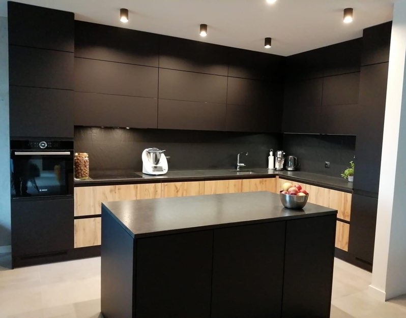 Nowoczesna, czarna kuchnia z drewnianymi akcentami, wyspą i oświetleniem punktowym; na blacie misa z owocami i robot kuchenny.