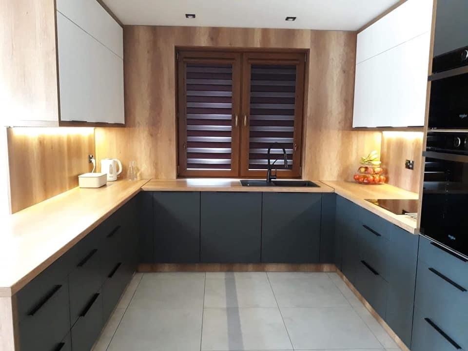 Nowoczesna kuchnia w kształcie litery U z drewnianymi blatami, ciemnymi szafkami i białymi szafkami wiszącymi, oświetlona listwami LED pod szafkami, z oknem z żaluzjami poziomymi i dekoracyjną...