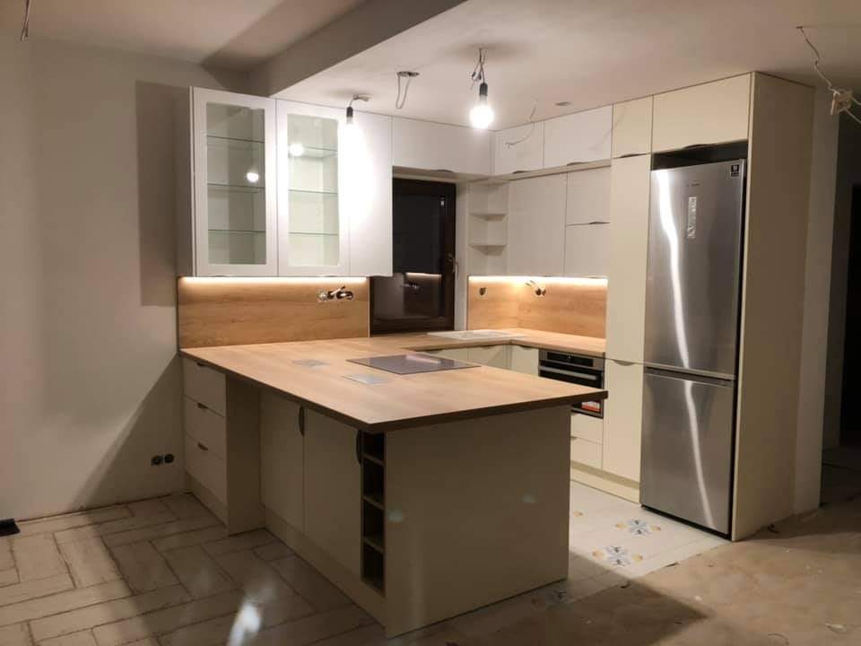 Jasna kuchnia z wyspą w trakcie montażu, widoczne niedokończone podłogi i wiszące kable, oświetlenie LED pod szafkami, lodówka side-by-side.