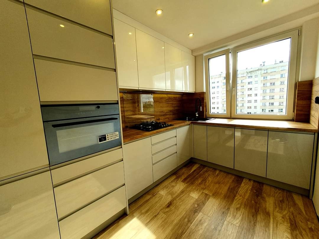 Jasna kuchnia na wymiar z drewnianym blatem i panelami, widok na budynek mieszkalny przez okno.