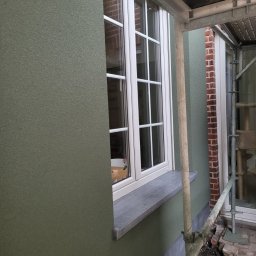 Tomek - Zielona elewacja z oknem po nałożeniu strukturalnego tynku, widoczne rusztowanie i ceglana ściana w tle. Detal wykończenia elewacji.