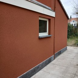 Tomek - Elewacja domu w odcieniach terakoty z oknem i szarą podmurówką, widok z boku na tle brukowanego chodnika i roślinności.