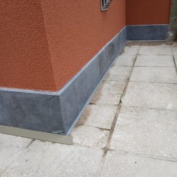 Tomek - Wykończona elewacja budynku z wyraźną, strukturalną fakturą pomarańczowego tynku i ciemnoszarą podmurówką. Widoczny fragment chodnika z płyt betonowych i małe okno z kratą.