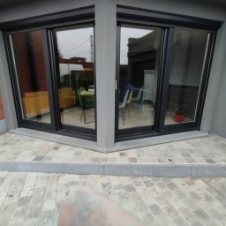 Tomek - Nowoczesne okna przesuwne z czarnymi ramami, umieszczone w narożniku budynku o szarym wykończeniu, z widokiem na wnętrze i fragmentem brukowanej nawierzchni.