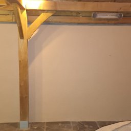 Tomek - Gładka, beżowa ściana wewnętrzna pod drewnianą konstrukcją dachu z widocznym grzejnikiem, wykończona płytkami na podłodze. W rogu widoczna drewniana boazeria.