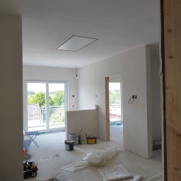 Tomek - Wnętrze w trakcie prac wykończeniowych: świeżo pomalowane ściany na biało, widoczne narzędzia malarskie, poziomica oparta o ścianę, okno balkonowe z widokiem na zieleń.