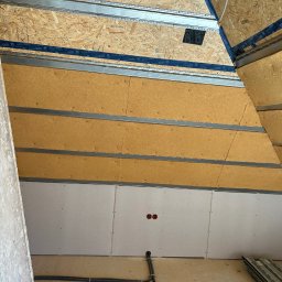 Tomek - Skośny sufit poddasza w trakcie zabudowy: płyty gipsowe, izolacja, profile metalowe, instalacja elektryczna. Widoczne materiały budowlane i etap prac wykończeniowych.