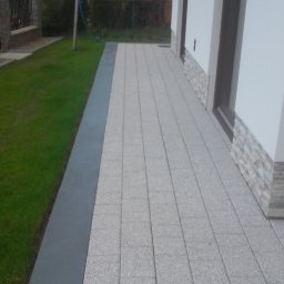 Tomek - Ułożona kostka brukowa przy budynku z trawnikiem, widok z góry. Szara kostka w regularnym wzorze, obrzeże z szarego betonu.