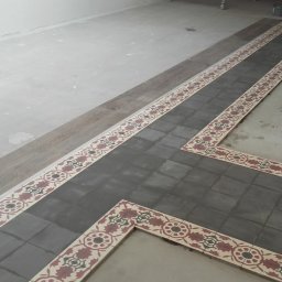 Tomek - Fragment podłogi z ułożoną szarą terakotą i dekoracyjnym bordiurem z motywem kwiatowym, widoczny fragment surowej posadzki i puszki z farbą.