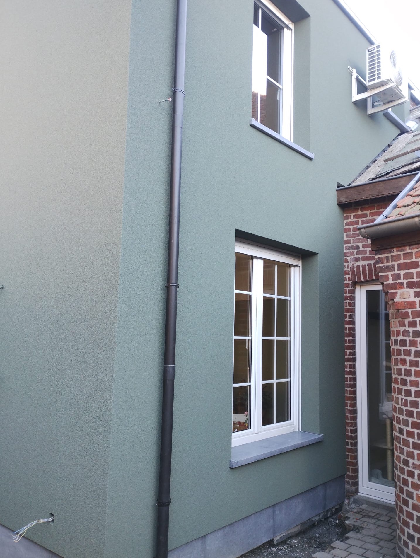 Elewacja budynku w odcieniach zieleni z widoczną rynną, oknami z białymi ramami i klimatyzatorem. Tynk ma delikatną fakturę. Widoczny fragment muru z czerwonej cegły.