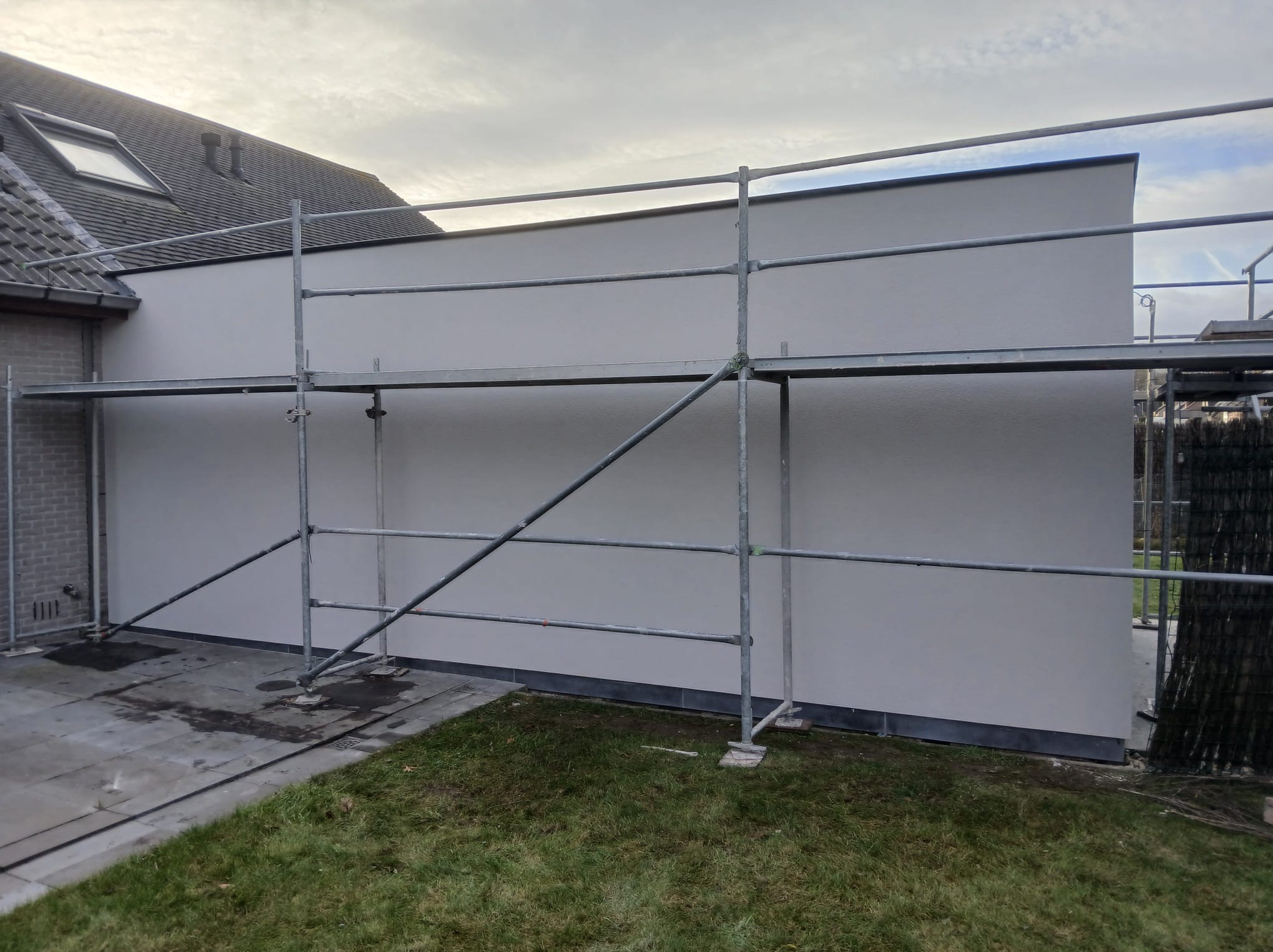 Świeżo otynkowana ściana domu jednorodzinnego z szarym wykończeniem, widoczna spod rusztowania na tle trawnika i pochmurnego nieba.