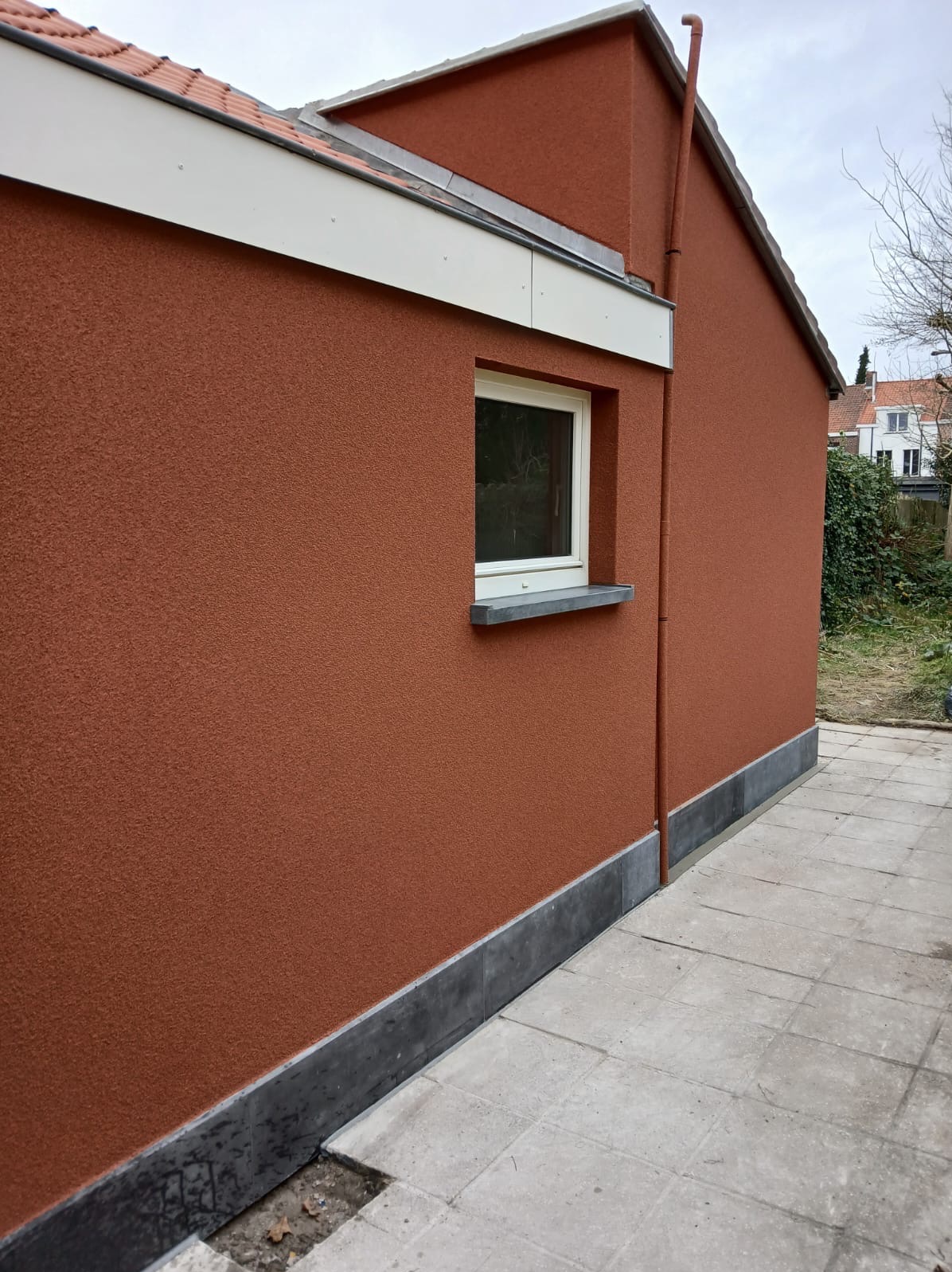 Elewacja domu w odcieniach terakoty z oknem i szarą podmurówką, widok z boku na tle brukowanego chodnika i roślinności.