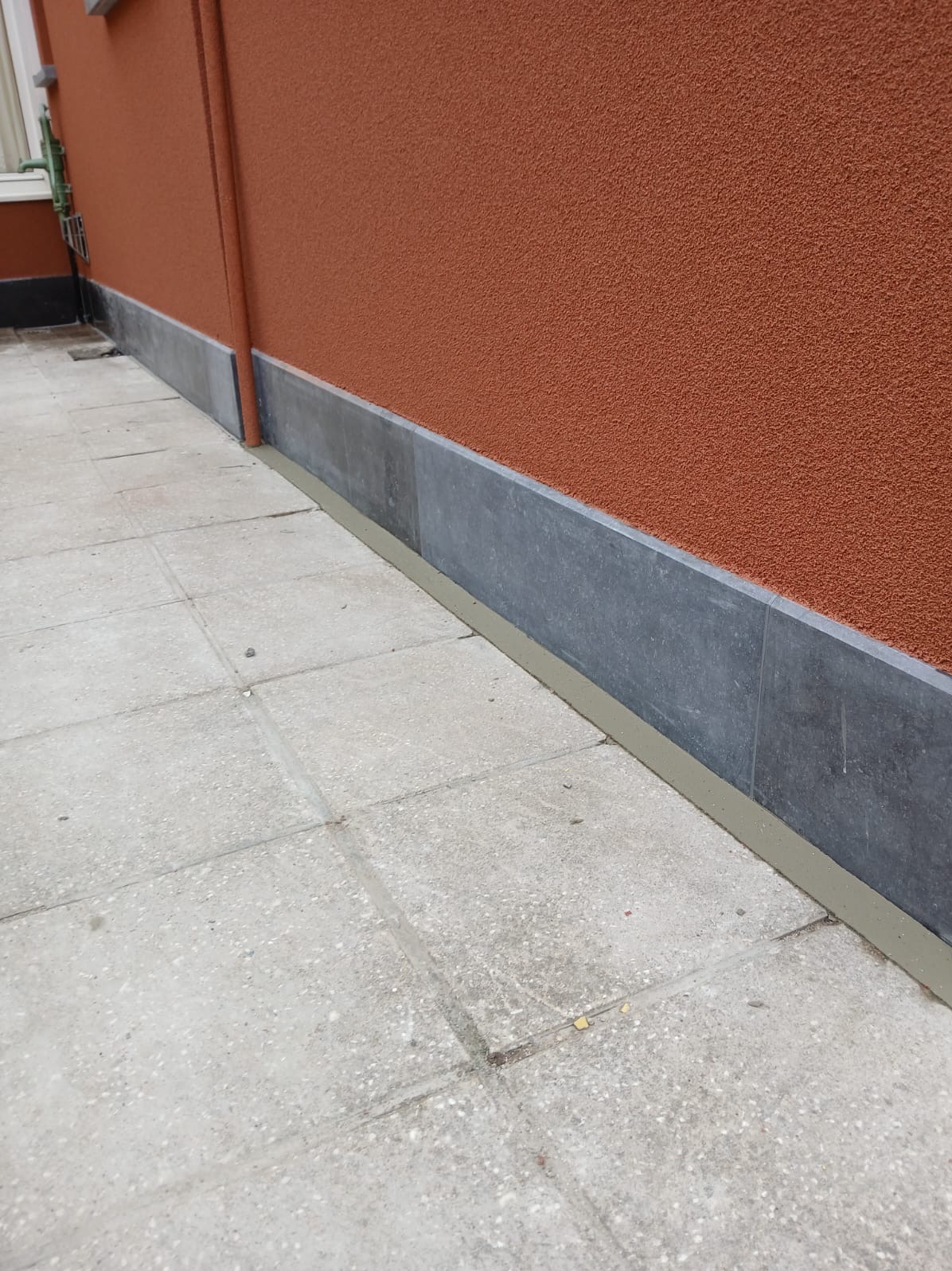 Teksturowana, pomarańczowa elewacja budynku z szarą, kamienną podmurówką i betonowym chodnikiem. Widoczna rynna spustowa.