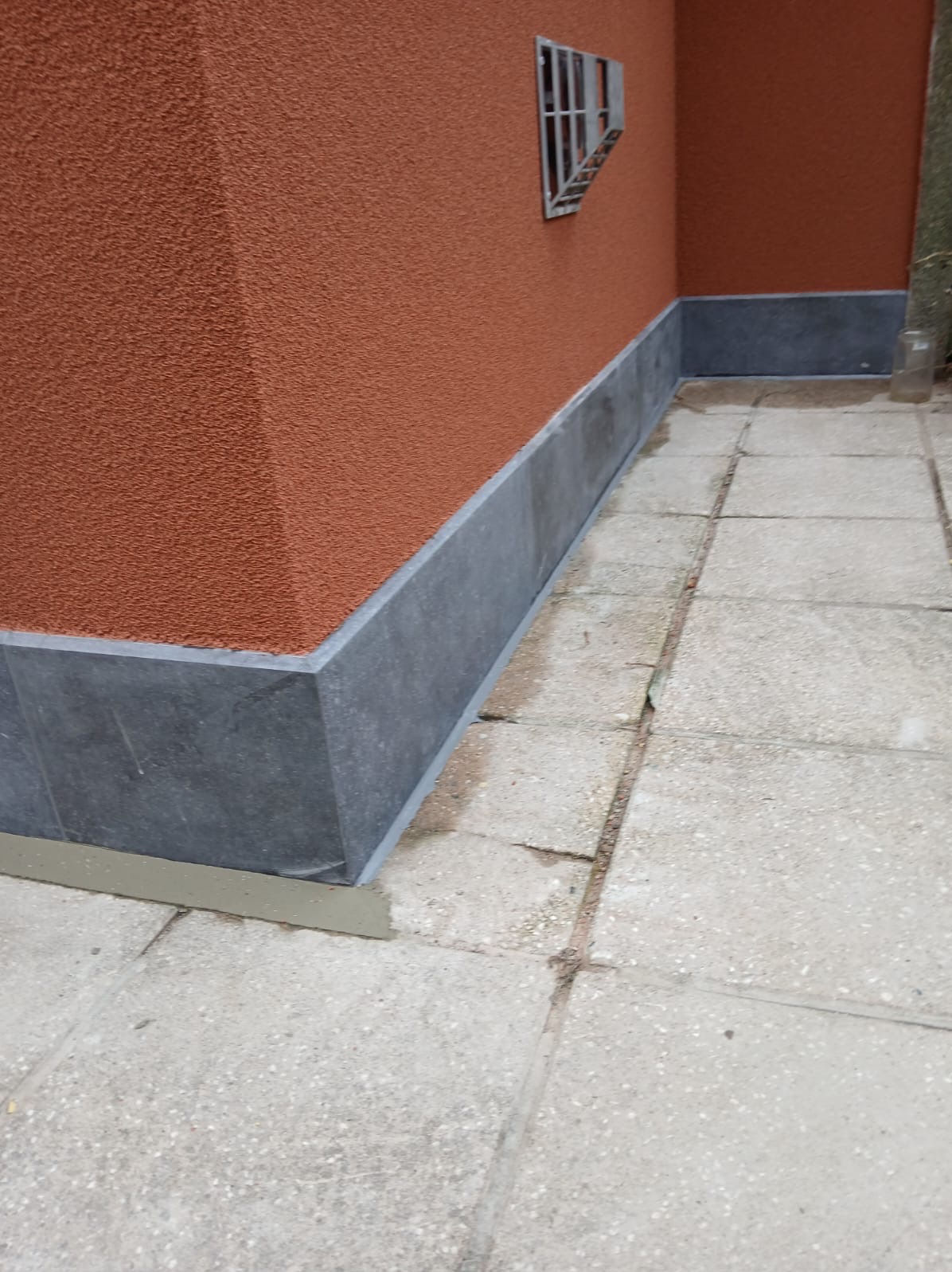 Wykończona elewacja budynku z wyraźną, strukturalną fakturą pomarańczowego tynku i ciemnoszarą podmurówką. Widoczny fragment chodnika z płyt betonowych i małe okno z kratą.