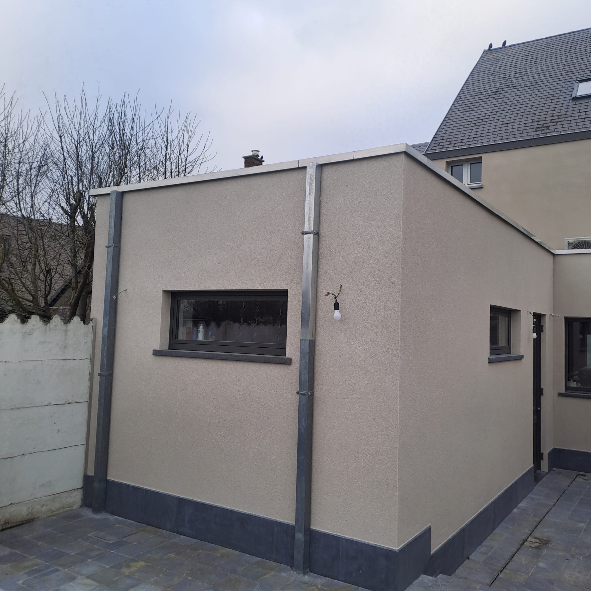 Elewacja budynku po ociepleniu, z widoczną strukturą tynku, oknami i rynnami. Detal: wisząca żarówka przy rynnie. Tło: budynek sąsiedni i drzewa bez liści.