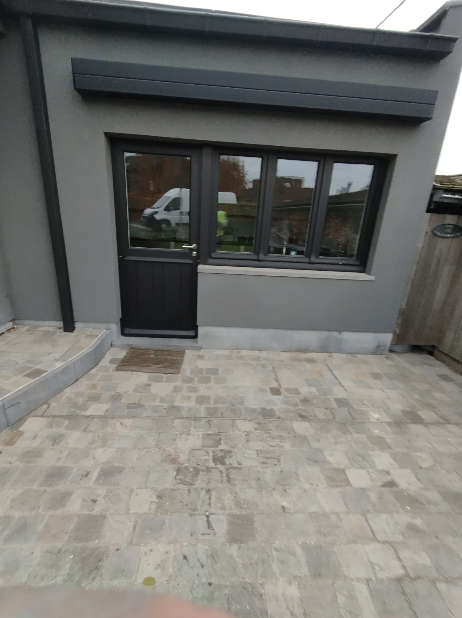 Szara elewacja budynku z czarnymi oknami i drzwiami, widoczne kamienne płytki na podłożu. W oknach odbija się biały samochód dostawczy.