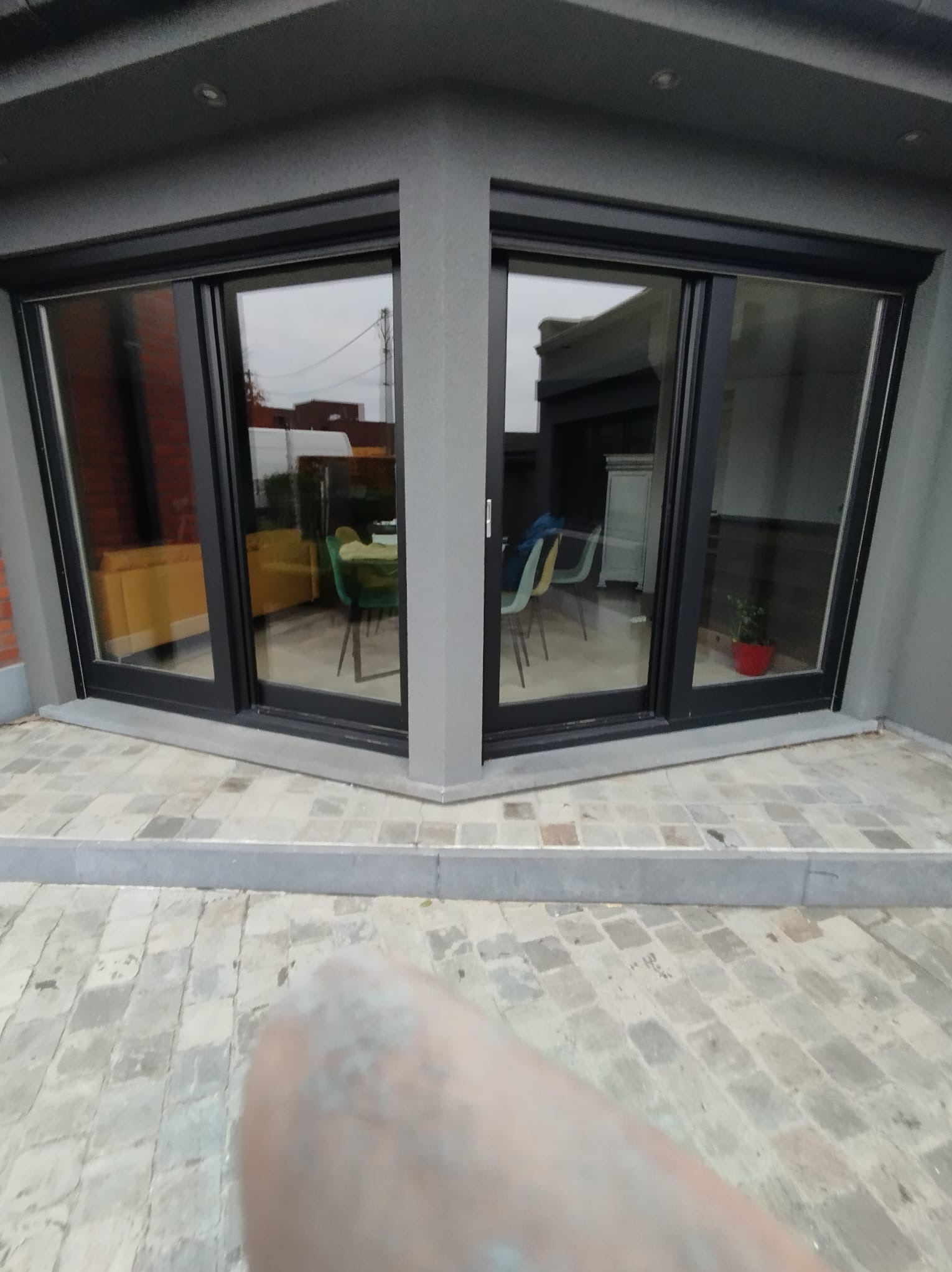 Nowoczesne okna przesuwne z czarnymi ramami, umieszczone w narożniku budynku o szarym wykończeniu, z widokiem na wnętrze i fragmentem brukowanej nawierzchni.
