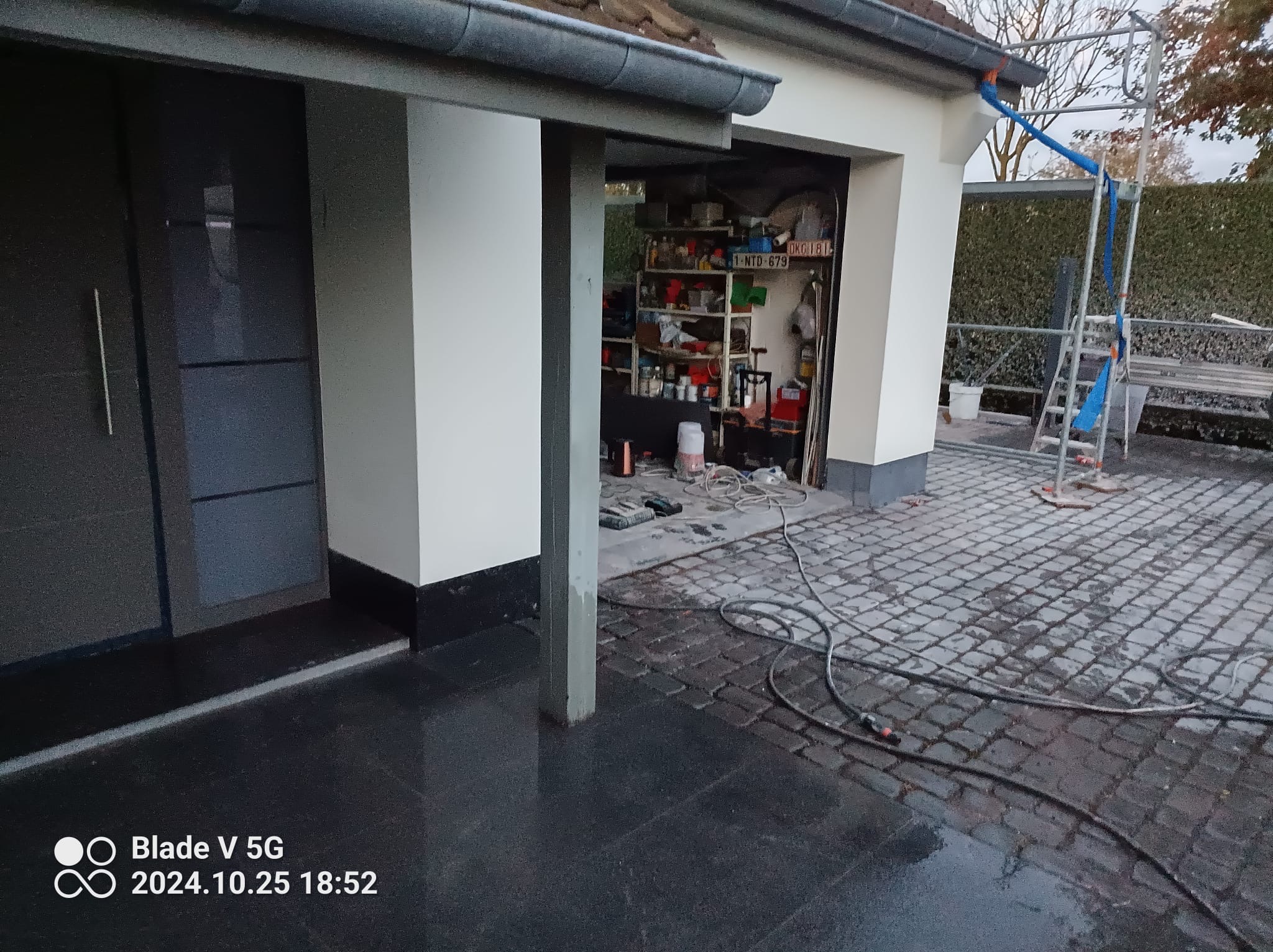 Odnowiona elewacja domu z szarymi detalami, widoczne rusztowanie i narzędzia, kostka brukowa na podjeździe. Prace wykończeniowe w toku, garaż otwarty.