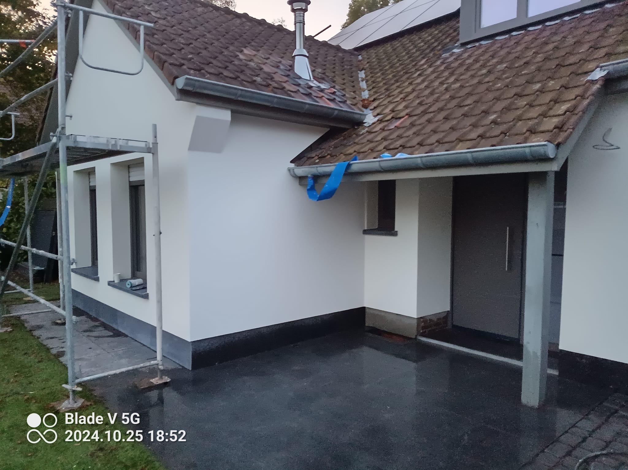 Biała elewacja domu po malowaniu, widoczne rusztowanie i fragment dachu z kominem. Świeżo pomalowana ściana i okna, ciemna podmurówka. Niebieska taśma zabezpieczająca rynnę.