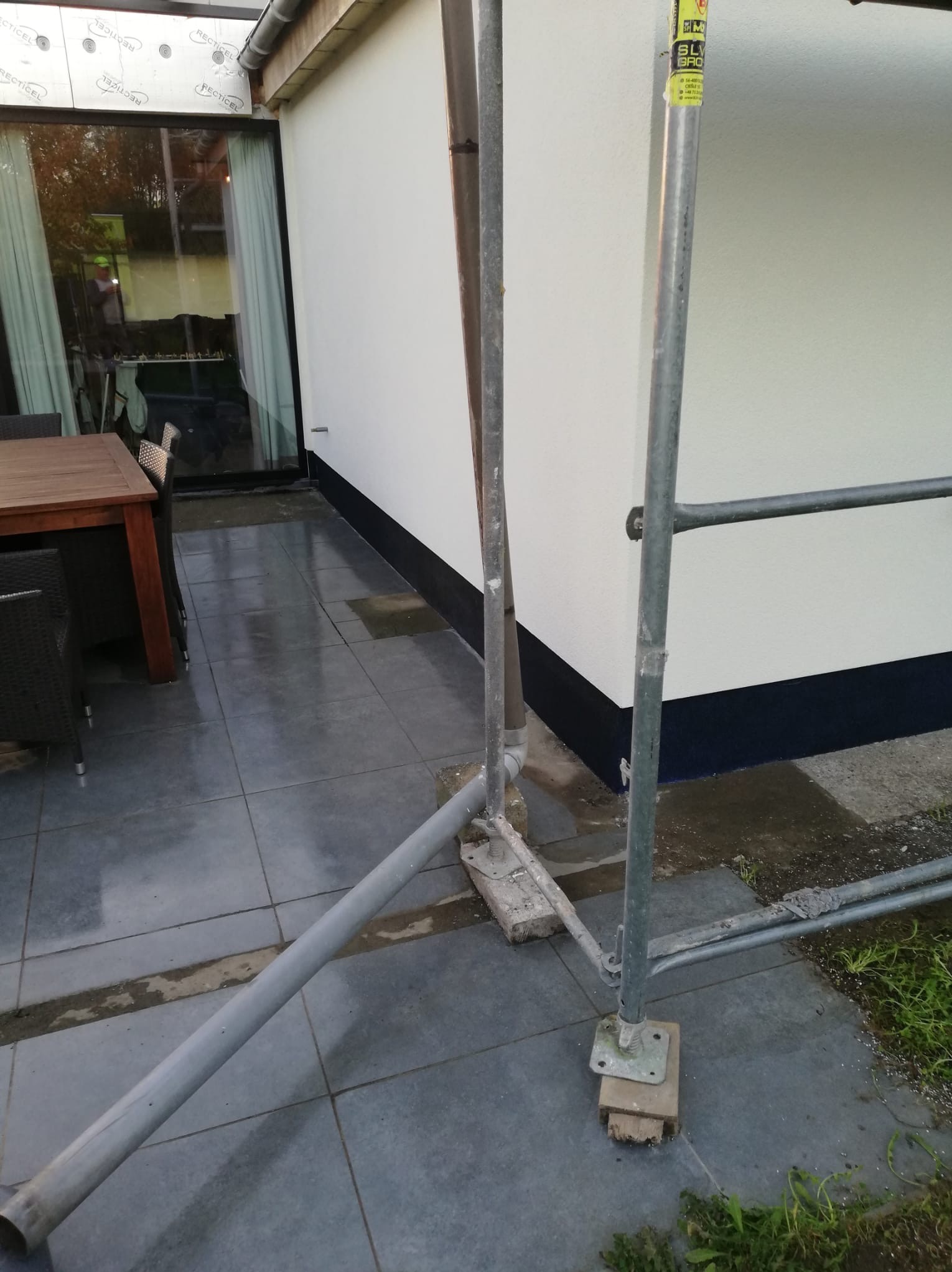 Ocieplona ściana budynku po malowaniu, z widocznymi rusztowaniami i fragmentem tarasu z meblami. Ściana w kolorze białym z czarnym pasem u dołu.