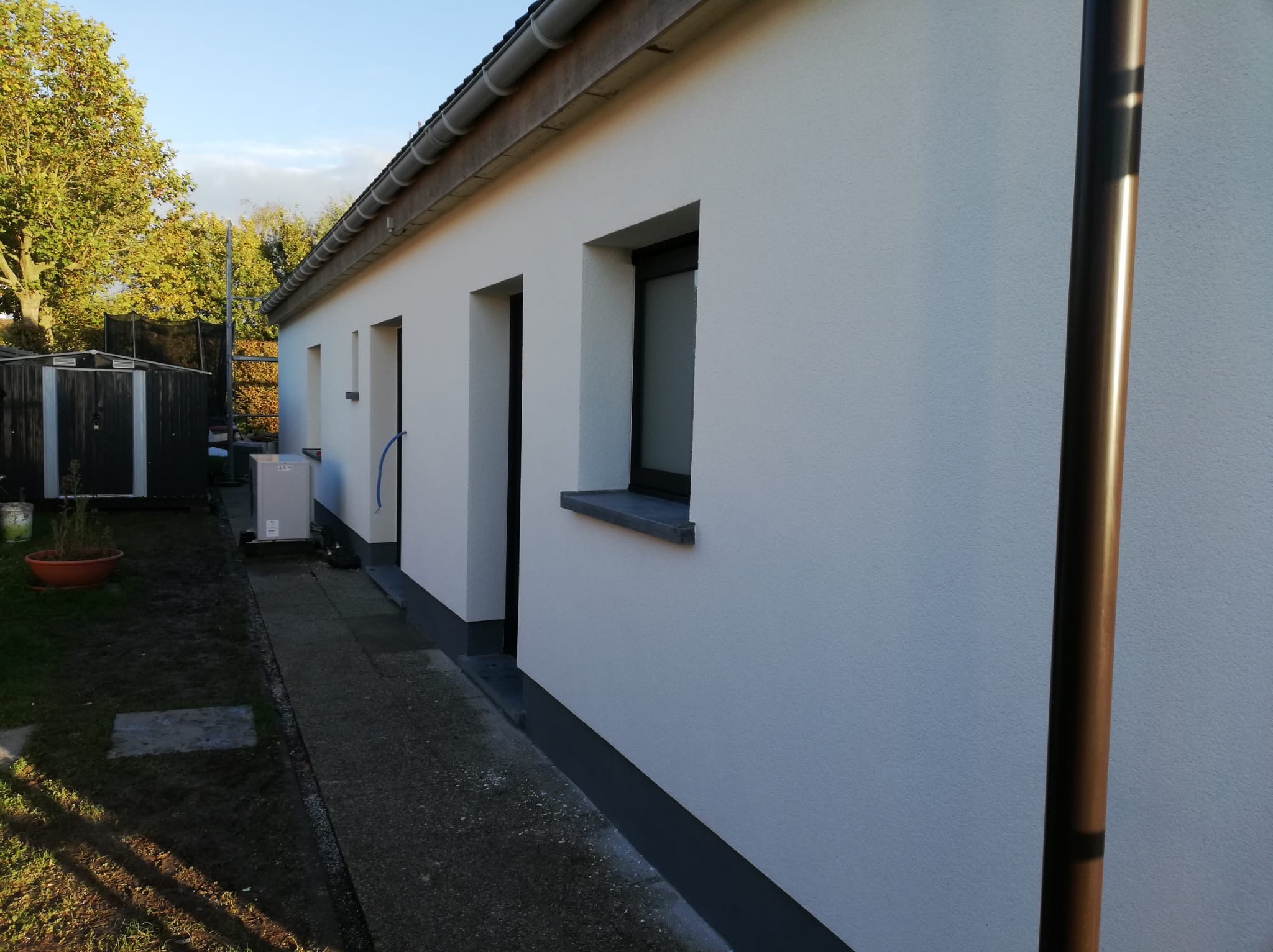 Ściana budynku po ociepleniu i pomalowaniu na biało. Widoczne okna z szarymi parapetami, brązowa rynna i fragment ogrodu z zielenią. Słoneczny dzień.