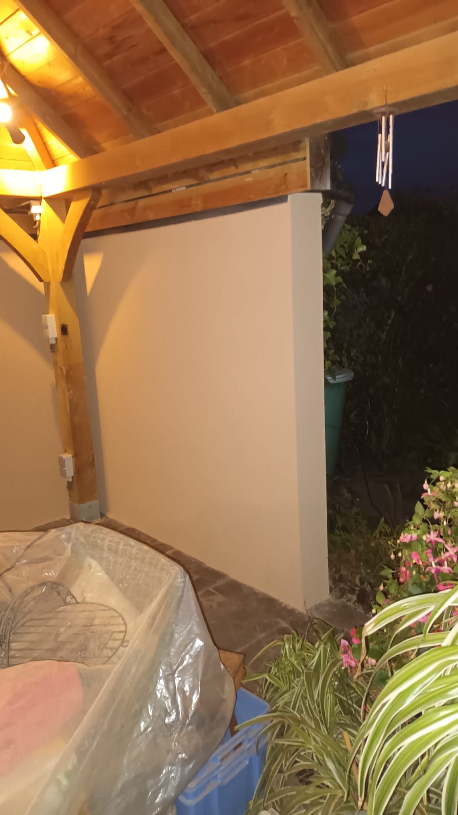 Świeżo pomalowana ściana zewnętrzna pod drewnianym zadaszeniem, z widocznymi narzędziami malarskimi zabezpieczonymi folią i zielenią w tle.