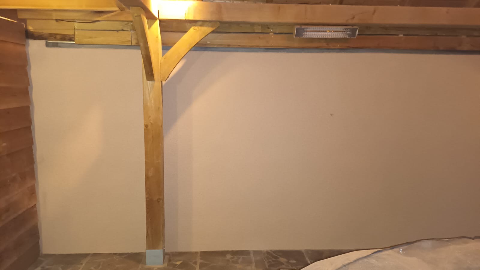 Gładka, beżowa ściana wewnętrzna pod drewnianą konstrukcją dachu z widocznym grzejnikiem, wykończona płytkami na podłodze. W rogu widoczna drewniana boazeria.