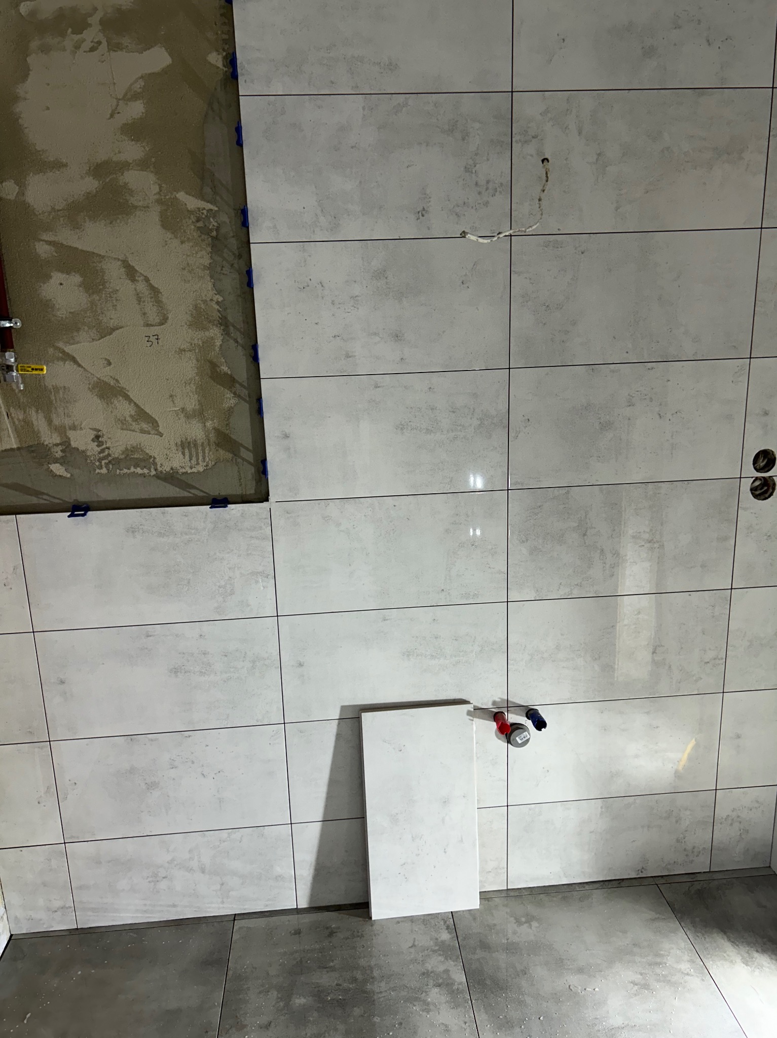 Ściana łazienki w trakcie wykańczania z szarymi płytkami imitującymi beton, widoczne fragmenty nieotynkowanej ściany i wystające rury.