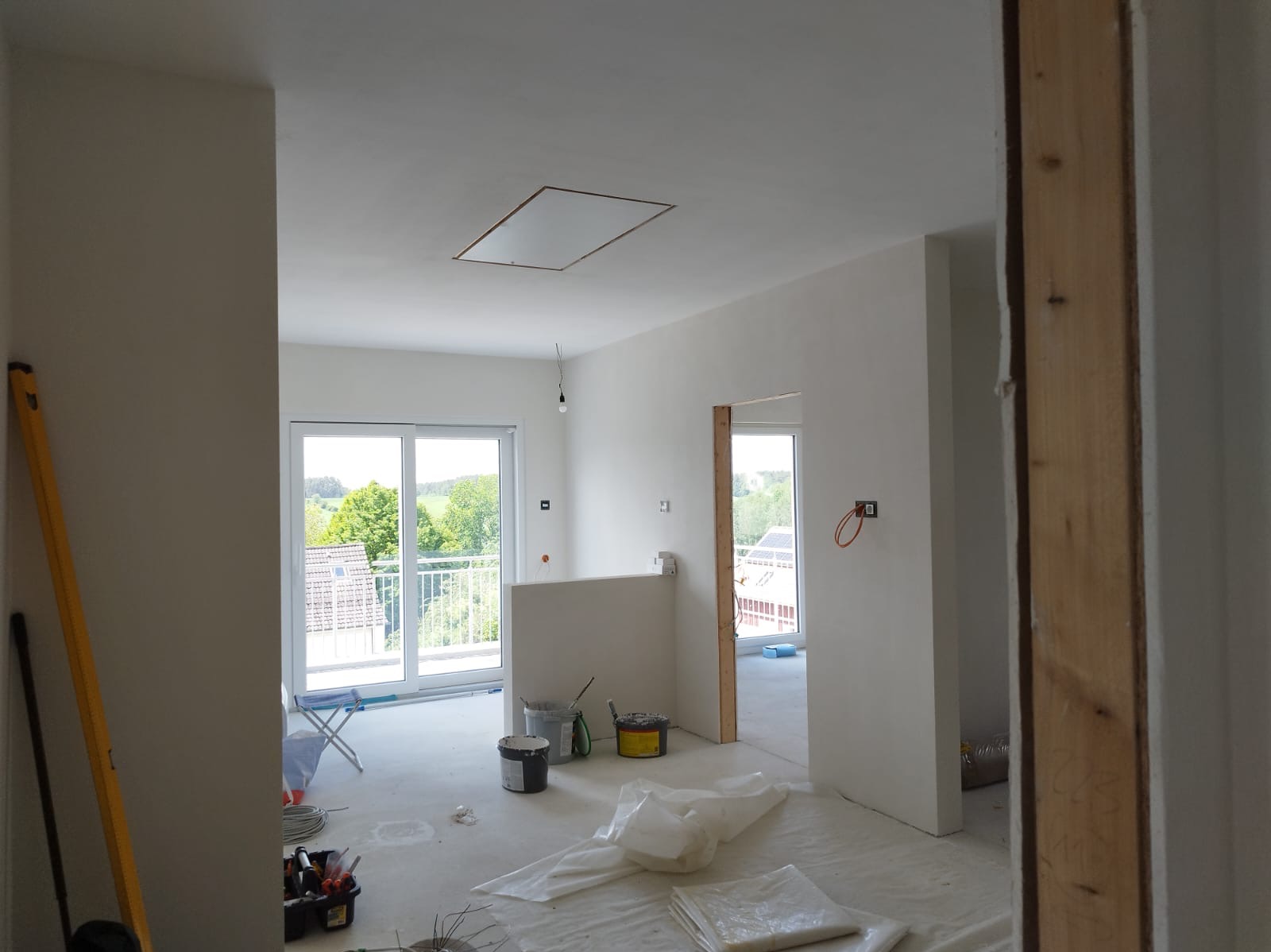 Wnętrze w trakcie prac wykończeniowych: świeżo pomalowane ściany na biało, widoczne narzędzia malarskie, poziomica oparta o ścianę, okno balkonowe z widokiem na zieleń.
