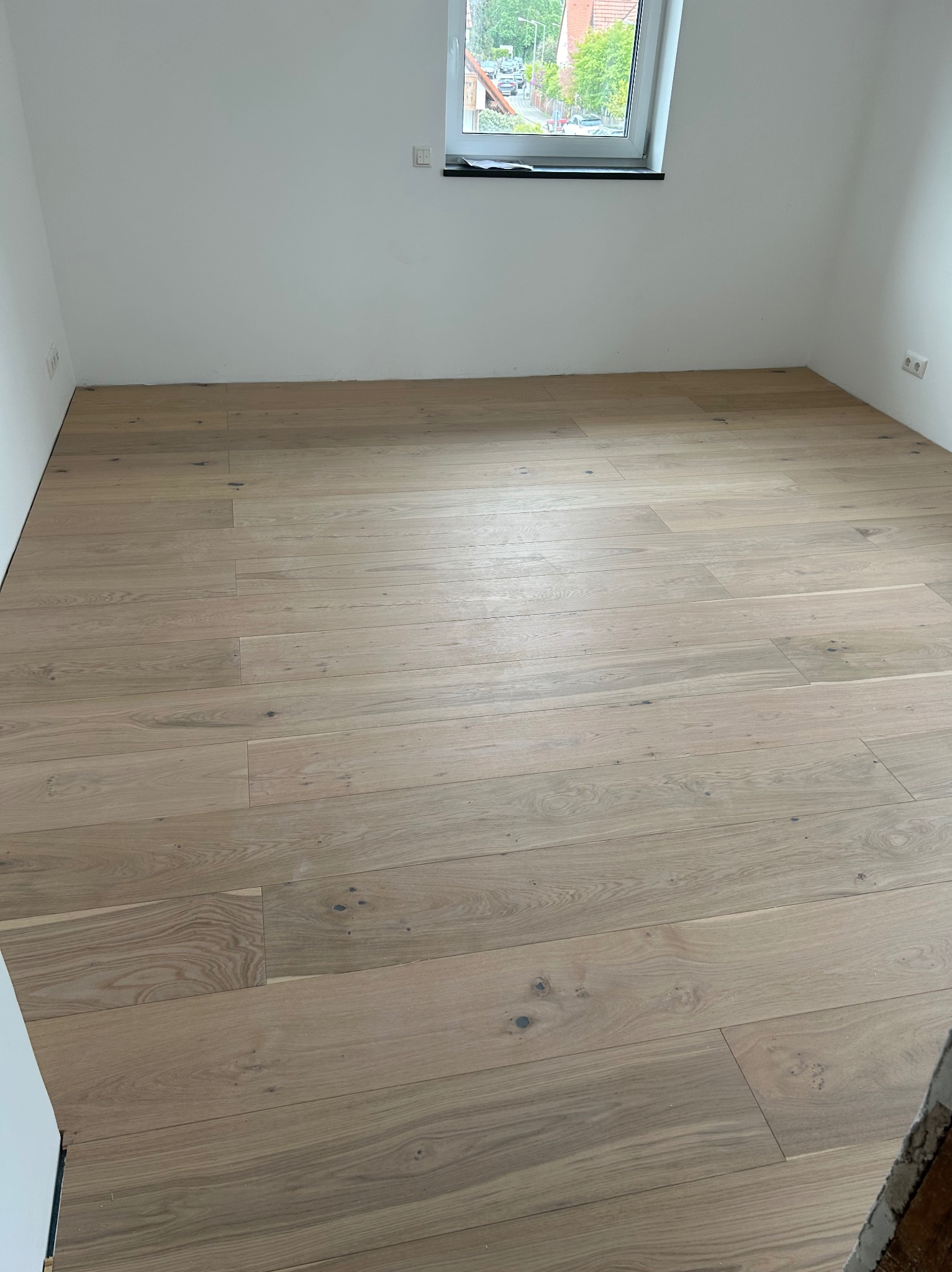 Jasne, drewniane panele podłogowe ułożone w pustym pokoju z białymi ścianami i oknem. Widoczne detale naturalnego drewna i subtelne usłojenie.