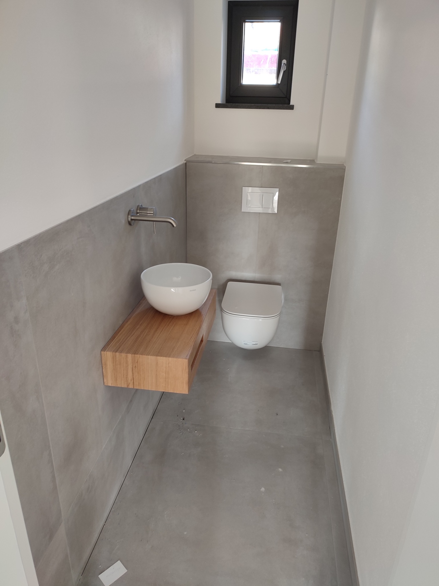 Nowoczesna łazienka z umywalką na drewnianym blacie, wiszącą toaletą i szarą okładziną ścienną w minimalistycznym stylu. Małe okno z czarną ramą w tle.