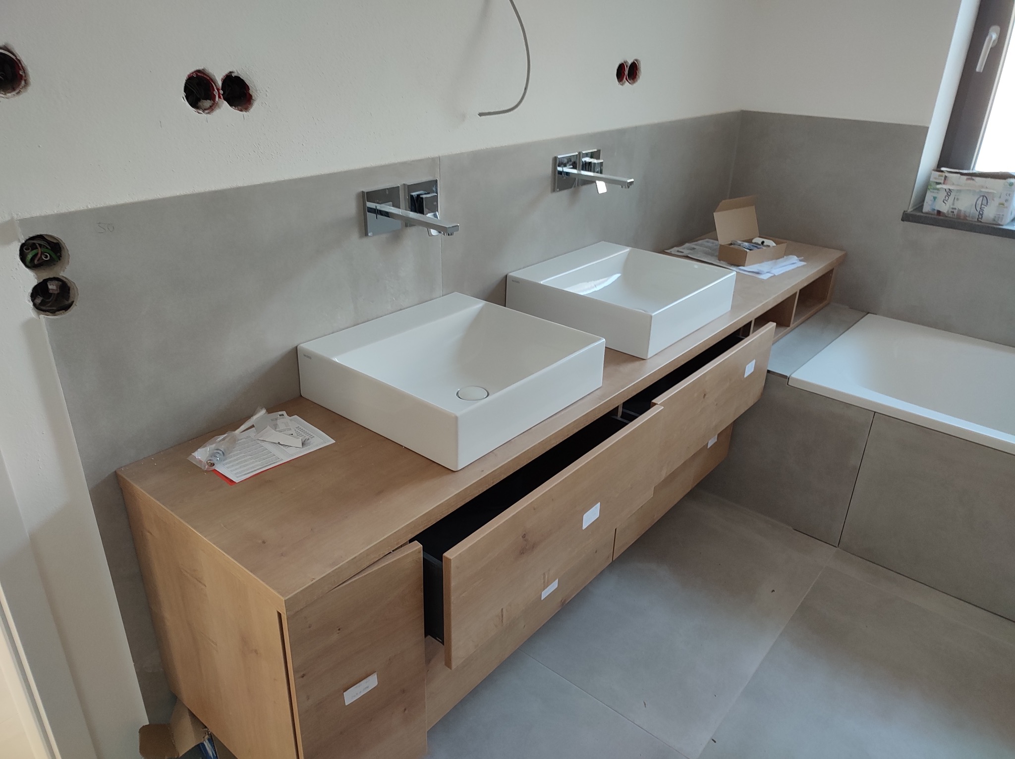 Dwie białe, kwadratowe umywalki nablatowe z nowoczesną armaturą na drewnianej szafce z szufladami w surowej łazience w trakcie wykończenia. Widoczne puszki elektryczne.