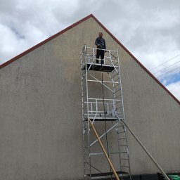 Paroclean Mobilna Myjnia Parowa - Pracownik na aluminiowym rusztowaniu fasadowym, przygotowujący się do mycia elewacji budynku o jasnoszarej, chropowatej strukturze, widoczny generator prądu na ziemi.