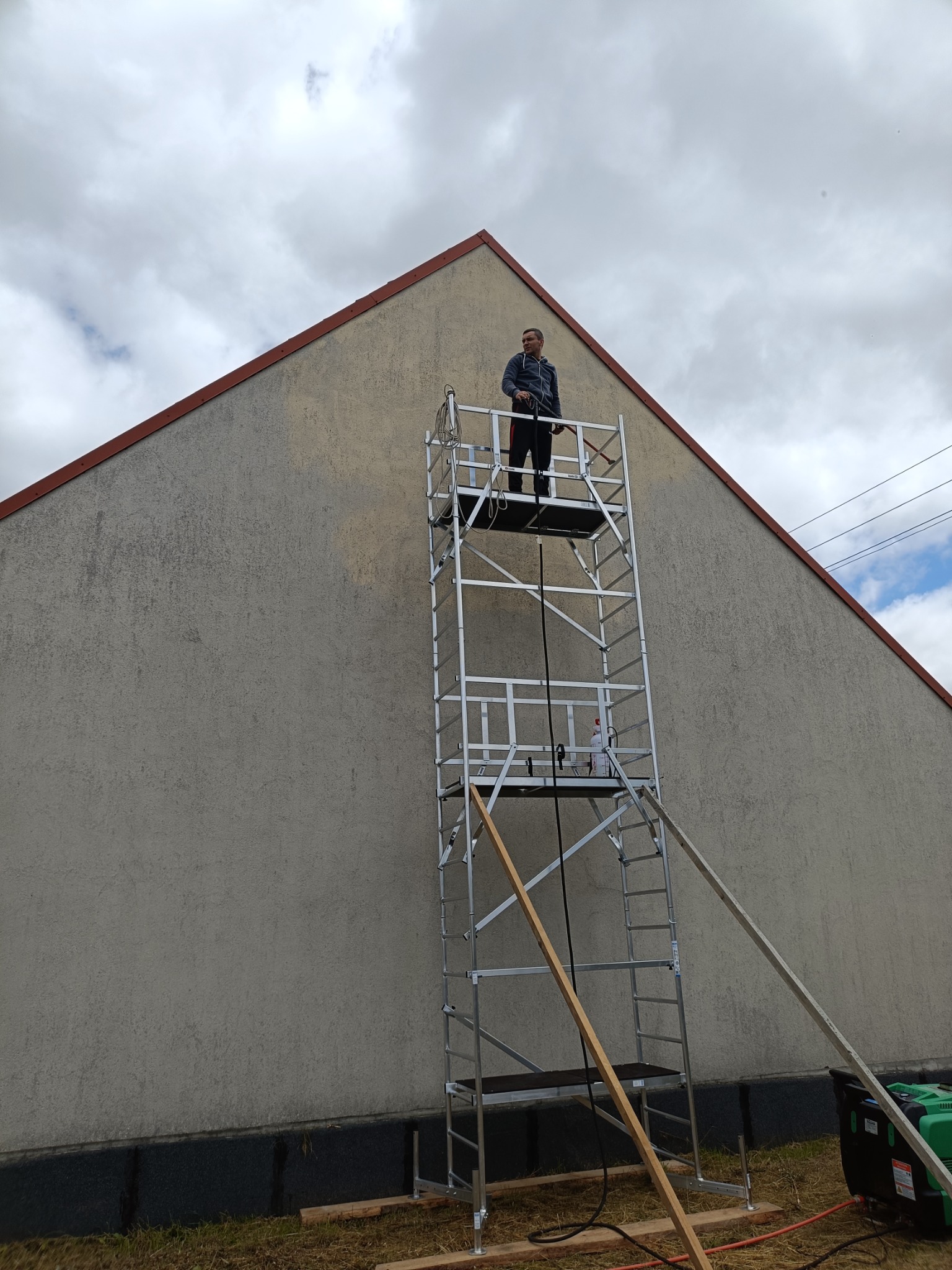 Pracownik na aluminiowym rusztowaniu fasadowym, przygotowujący się do mycia elewacji budynku o jasnoszarej, chropowatej strukturze, widoczny generator prądu na ziemi.