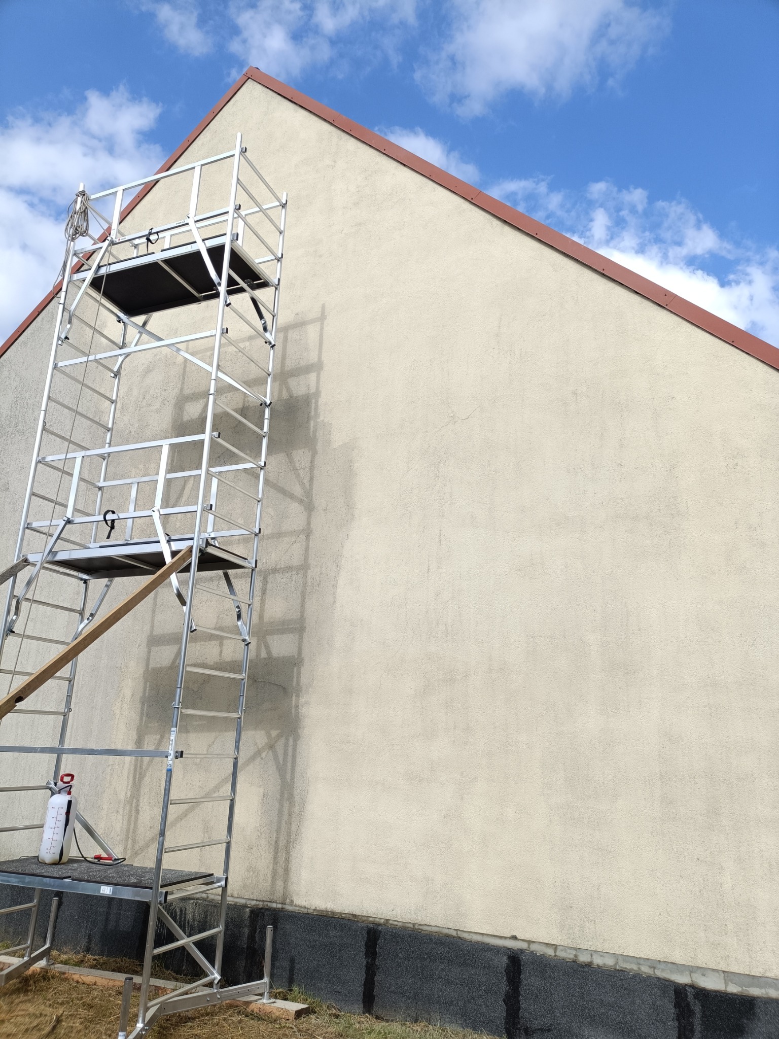 Ściana budynku z beżową elewacją, częściowo umyta, z widocznym rusztowaniem aluminiowym i urządzeniem ciśnieniowym na podeście, z ciemną, wilgotną podstawą elewacji.
