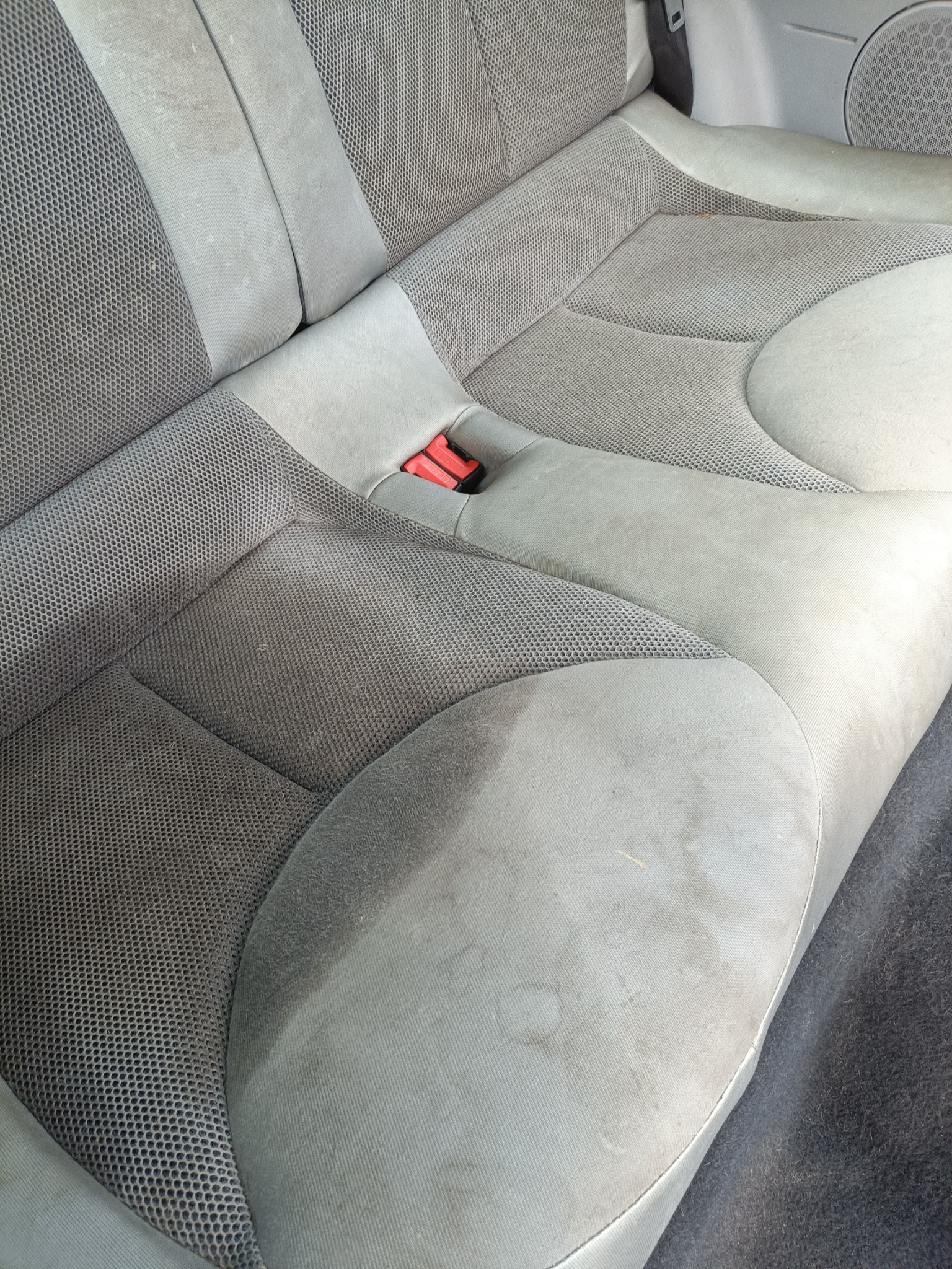 Brudna, szara tapicerka tylnej kanapy samochodowej z plamami i zabrudzeniami, widoczne czerwone zapięcie pasów.