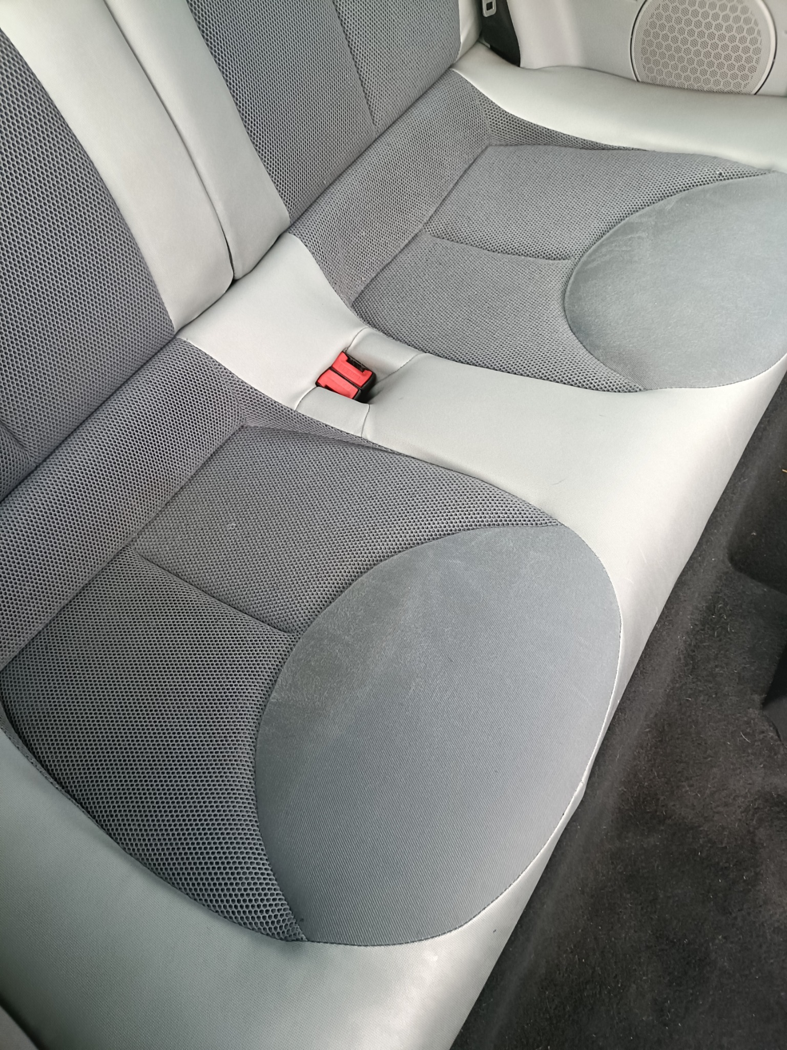 Szare, dwuosobowe siedzenie samochodowe z elementami z siatki i czerwonym zapięciem pasa bezpieczeństwa, widoczne ślady użytkowania.
