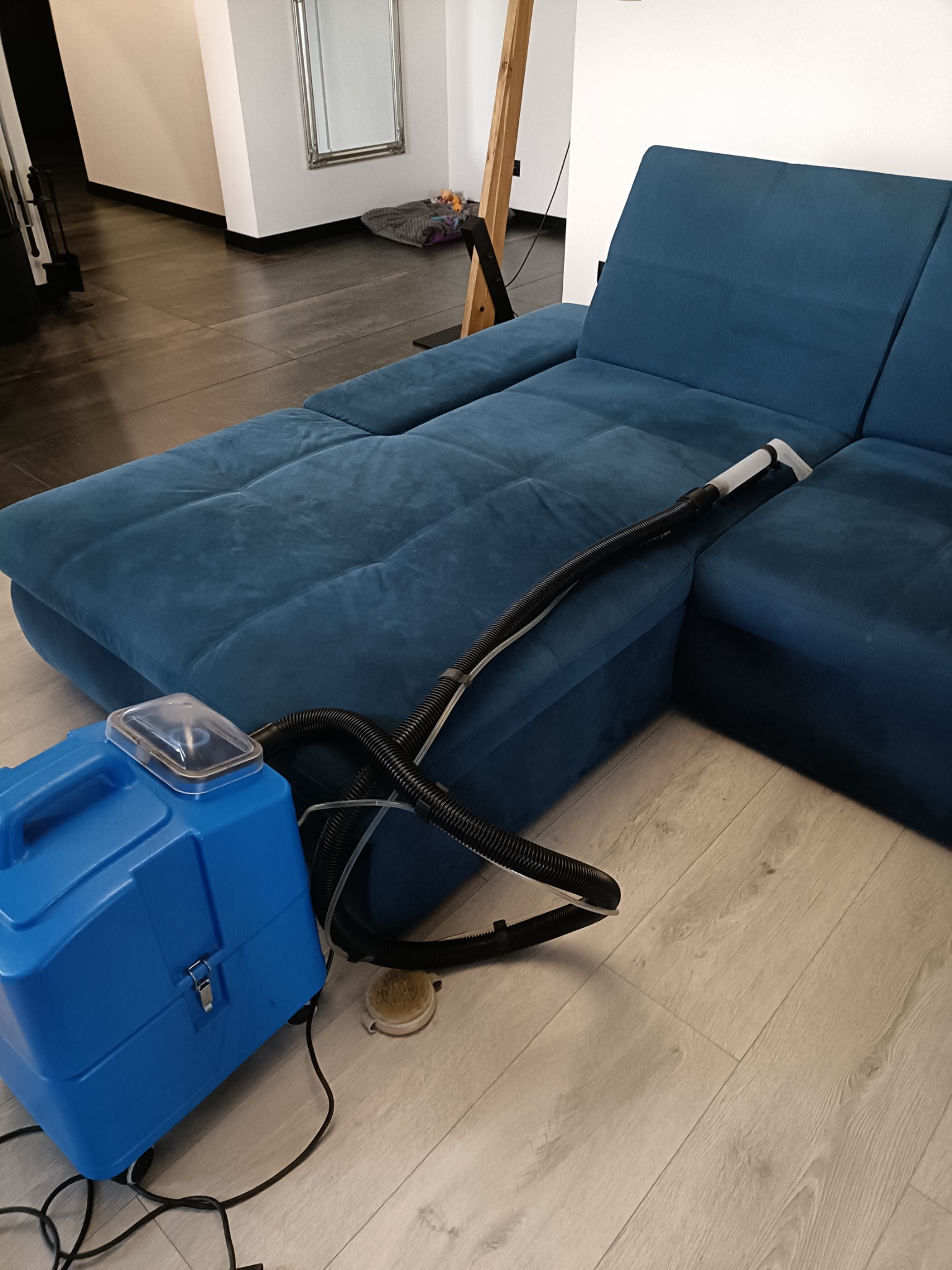 Niebieska narożna kanapa z welurową tapicerką czyszczona za pomocą niebieskiego odkurzacza piorącego z przezroczystym zbiornikiem na wodę, widoczna dysza czyszcząca na siedzisku.