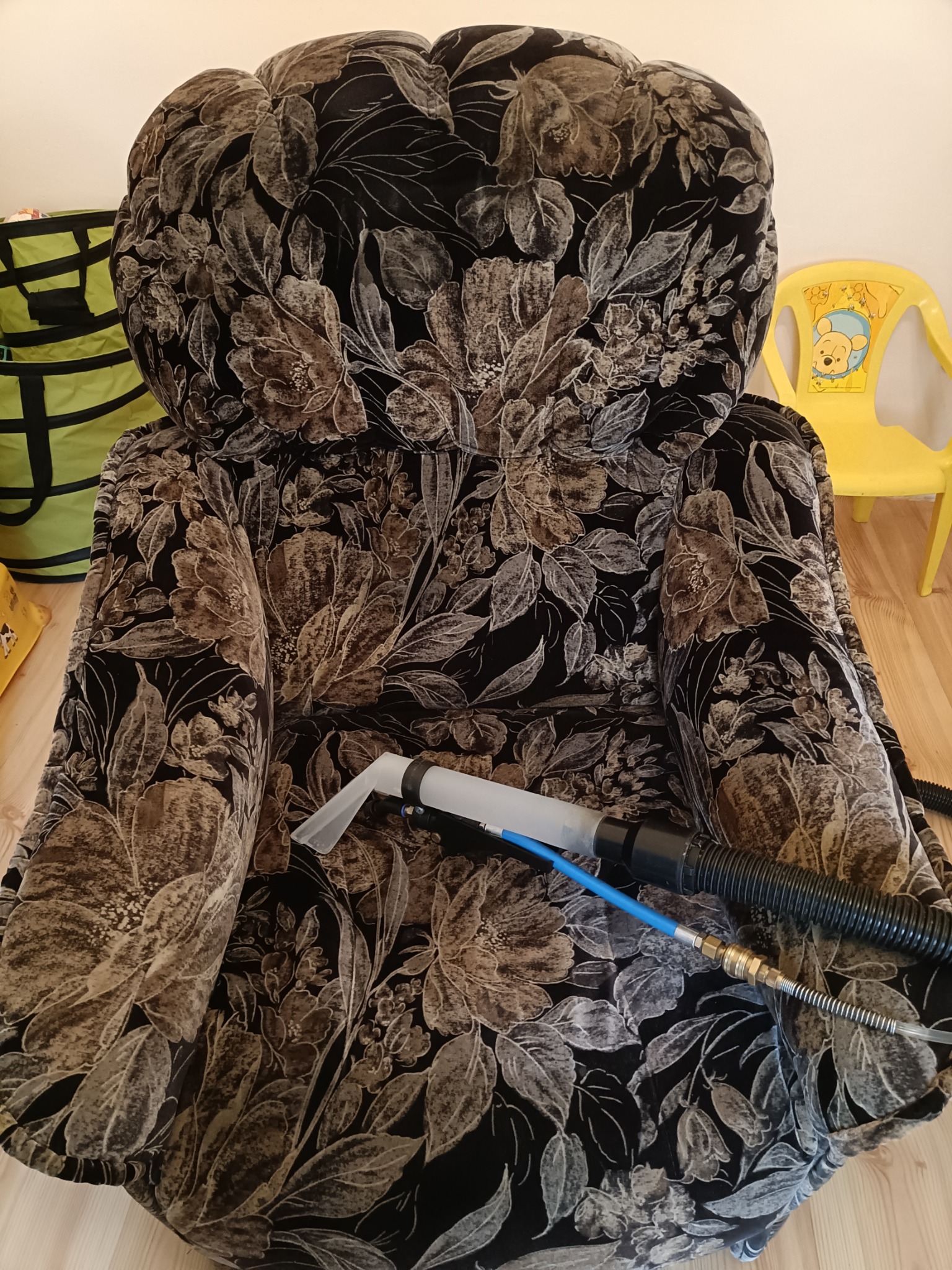 Fotel obity tkaniną w ciemne kwiaty, czyszczony za pomocą dyszy i węża odkurzacza piorącego; w tle widoczne zabawki.