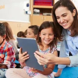 Uśmiechnięta nauczycielka pomaga dziewczynce z tabletem na tle grupy dzieci w klasie.