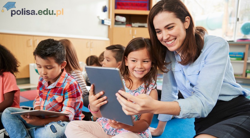 Uśmiechnięta nauczycielka pomaga dziewczynce z tabletem na tle grupy dzieci w klasie.