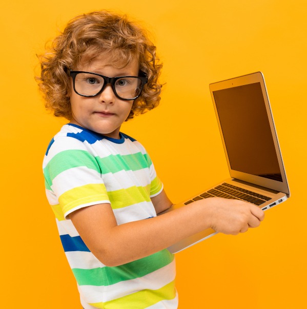 Chłopiec w okularach trzyma otwarty laptop na żółtym tle, ubrany w koszulkę w paski.